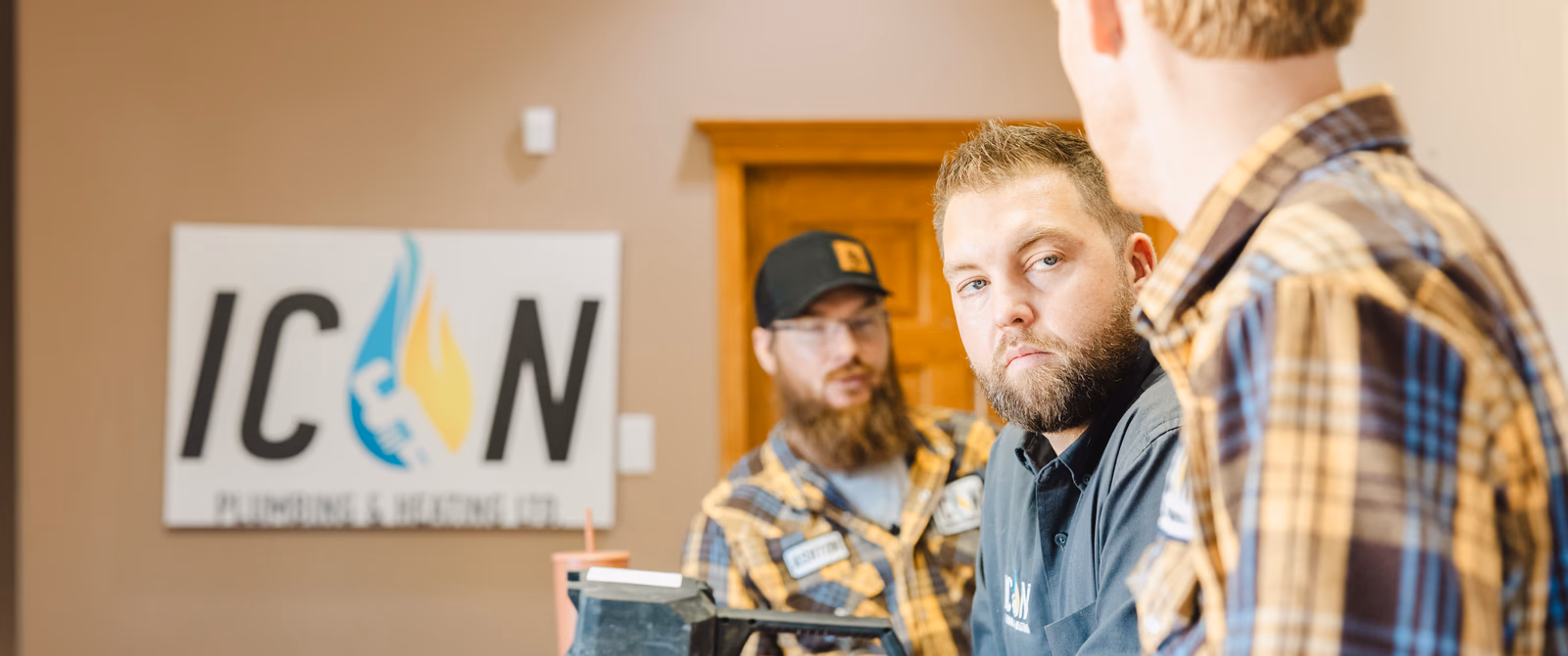 Two bearded men from ICON Plumbing & Heating Ltd. discussing work in an office with the company logo on the wall.