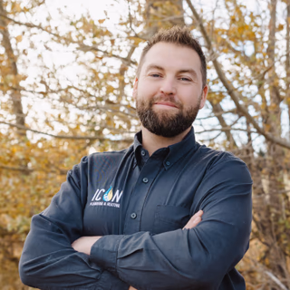 Professional plumber with a beard wearing a dark shirt with the Icon Plumbing & Heating logo, standing outdoors in autumn.