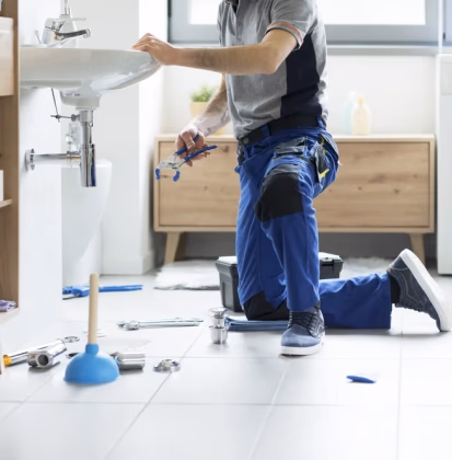 Plumber kneeling on a tiled floor while using pliers to repair a sink drain, with various tools and a plunger nearby.